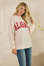 A woman wears a cream-colored, long-sleeved ALABAMA Cloudy Soft Knit Long Sleeve Top with a waffle-knit texture and “ALABAMA” in red varsity lettering across the chest.