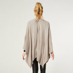 A woman with blond hair in a ponytail is shown from the back against a plain background, wearing the COCO + CARMEN Kiara Light Weight Wrap in light beige over black pants.