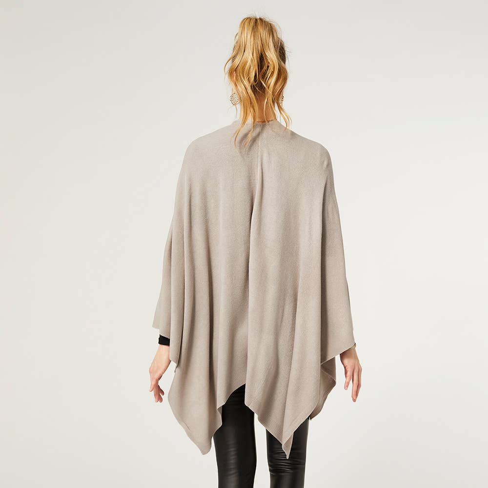 A woman with blond hair in a ponytail is shown from the back against a plain background, wearing the COCO + CARMEN Kiara Light Weight Wrap in light beige over black pants.