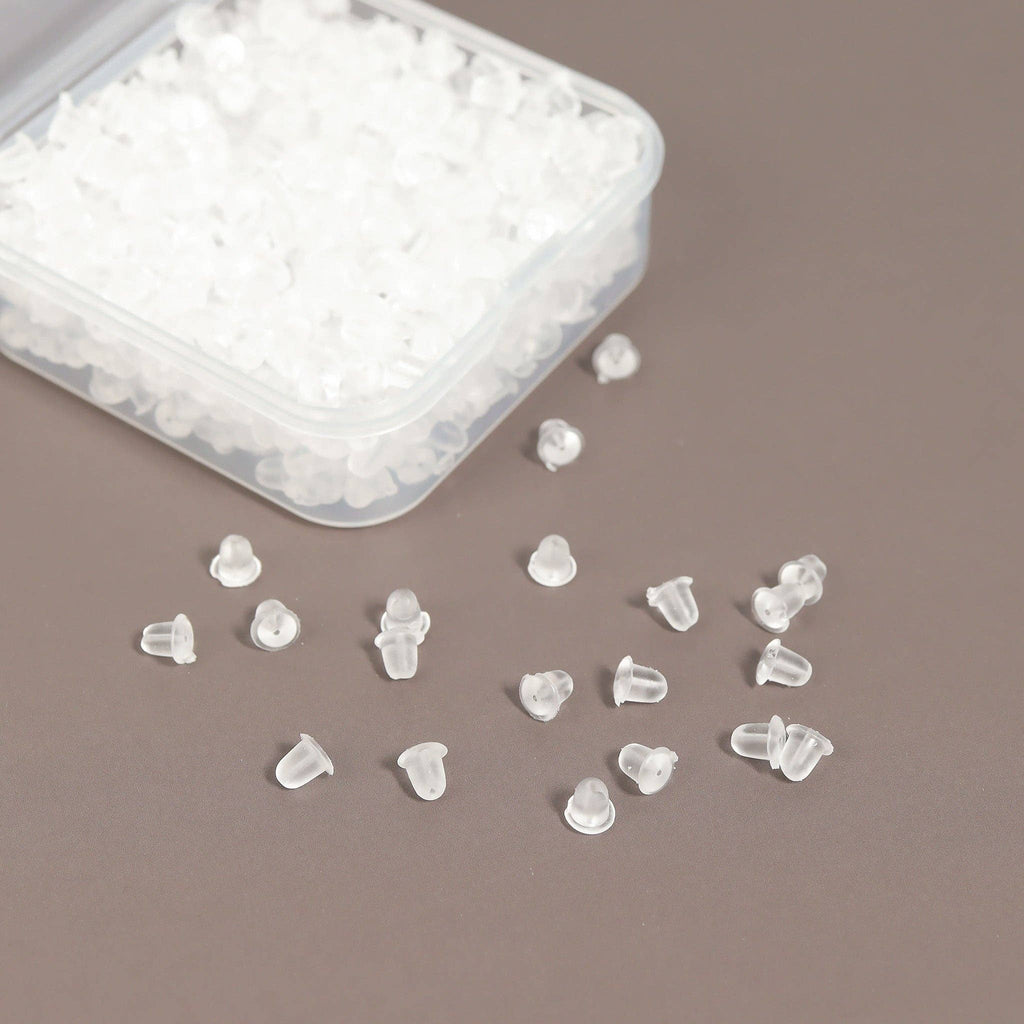A plastic container holds WJW Silicone Earring Backs (1 PAIR), with several secure earring backs scattered on a brown surface.