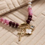 The Lilla Haven Textured Glass Bead Necklace with Charm Trio features pink and burgundy glass beads accented by gold-toned spacers, and is adorned with three gold charms—a cross, a clover, and a square gemstone—presented on beige textured fabric.