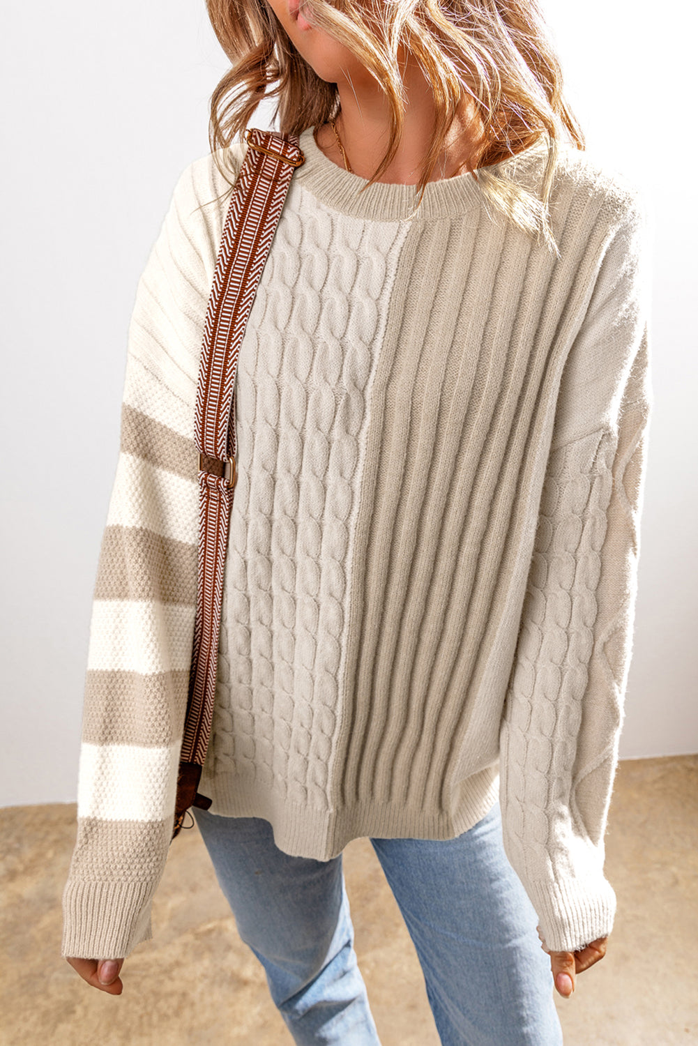 Someone wearing a Shewin Khaki Cable Knit Colorblock Crew Neck Drop Shoulder Sweater paired with blue jeans and a brown shoulder strap.