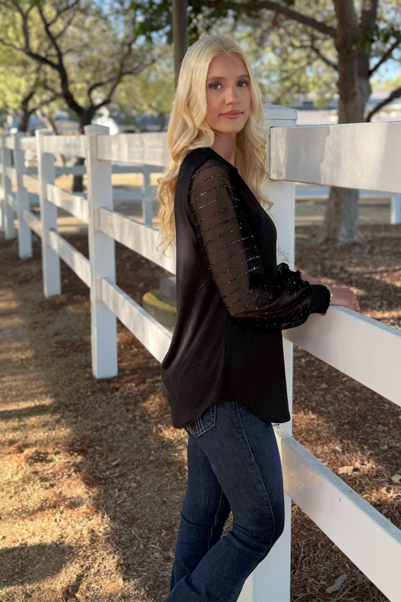 A woman with long blonde hair stands by a white wooden fence outdoors, wearing the 24-Hrs Fashion Sequin Sleeve Blouse and jeans, with trees and sunlight in the background.