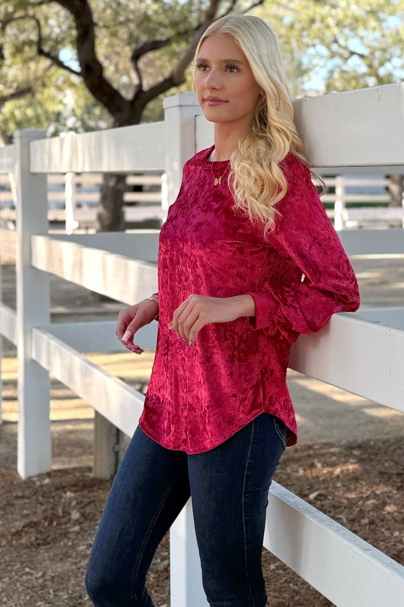 A woman stands outdoors by a white wooden fence, wearing the 24-Hrs Fashion Crushed Velvet Top with dark jeans and trees in the background.