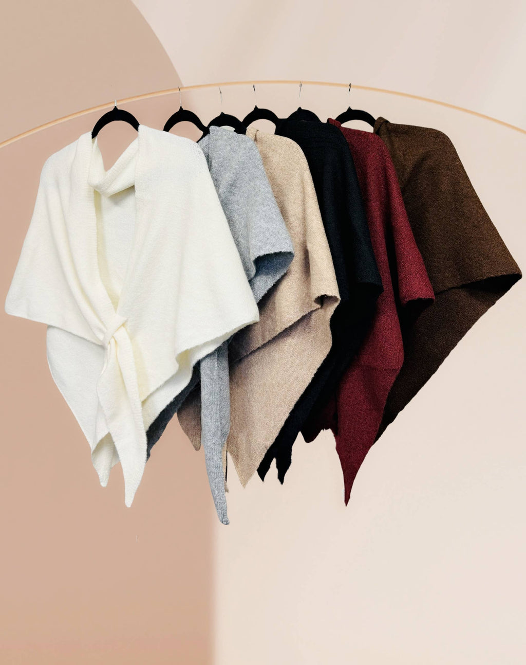 Six JC Sunny Fashion Cashmere-Blend Knit Triangle Shawls with cross-front design in white, gray, beige, taupe, burgundy, and brown hang on black hangers from a gold rack against a pale background.