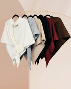 Six JC Sunny Fashion Cashmere-Blend Knit Triangle Shawls with cross-front design in white, gray, beige, taupe, burgundy, and brown hang on black hangers from a gold rack against a pale background.