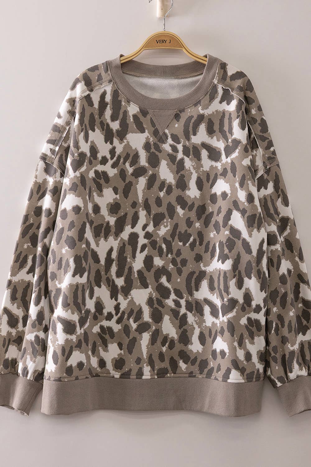 The Leopard Printed Crew Neck Sweatshirt by very J features a stylish gray and beige leopard print, long sleeves, and ribbed cuffs, hem, and neckline. Displayed on a wooden hanger against a light-colored wall, its relaxed fit offers effortless style.