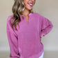 A woman in a Shewin Notched Neck Exposed Seam Drop Shoulder Sweatshirt, colored pink, and white pants is smiling against a plain beige background.