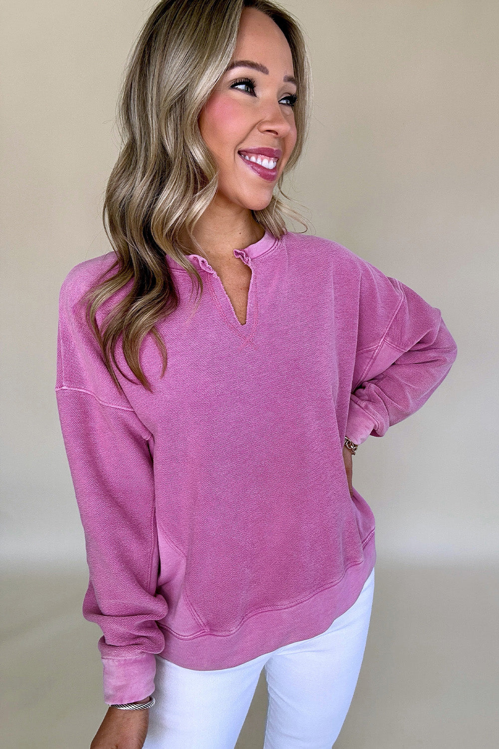 A woman in a Shewin Notched Neck Exposed Seam Drop Shoulder Sweatshirt, colored pink, and white pants is smiling against a plain beige background.