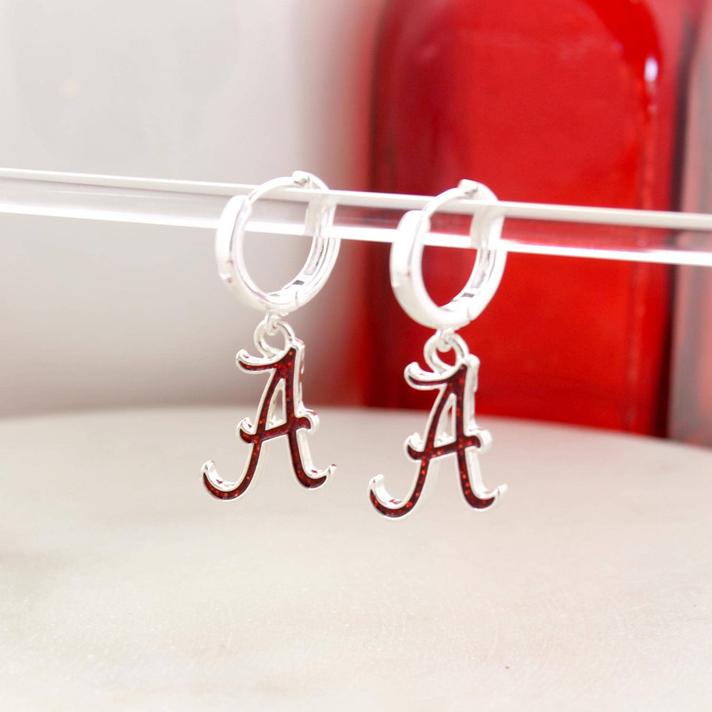Seasons Jewelry Alabama Glitter Enamel Logo Huggie Hoop Earrings feature silver huggie hoops with dangling red and white Alabama “A” logo charms accented by crimson glitter enamel, shown on a clear rod with a red object in the background.