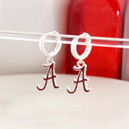 Seasons Jewelry Alabama Glitter Enamel Logo Huggie Hoop Earrings feature silver huggie hoops with dangling red and white Alabama “A” logo charms accented by crimson glitter enamel, shown on a clear rod with a red object in the background.
