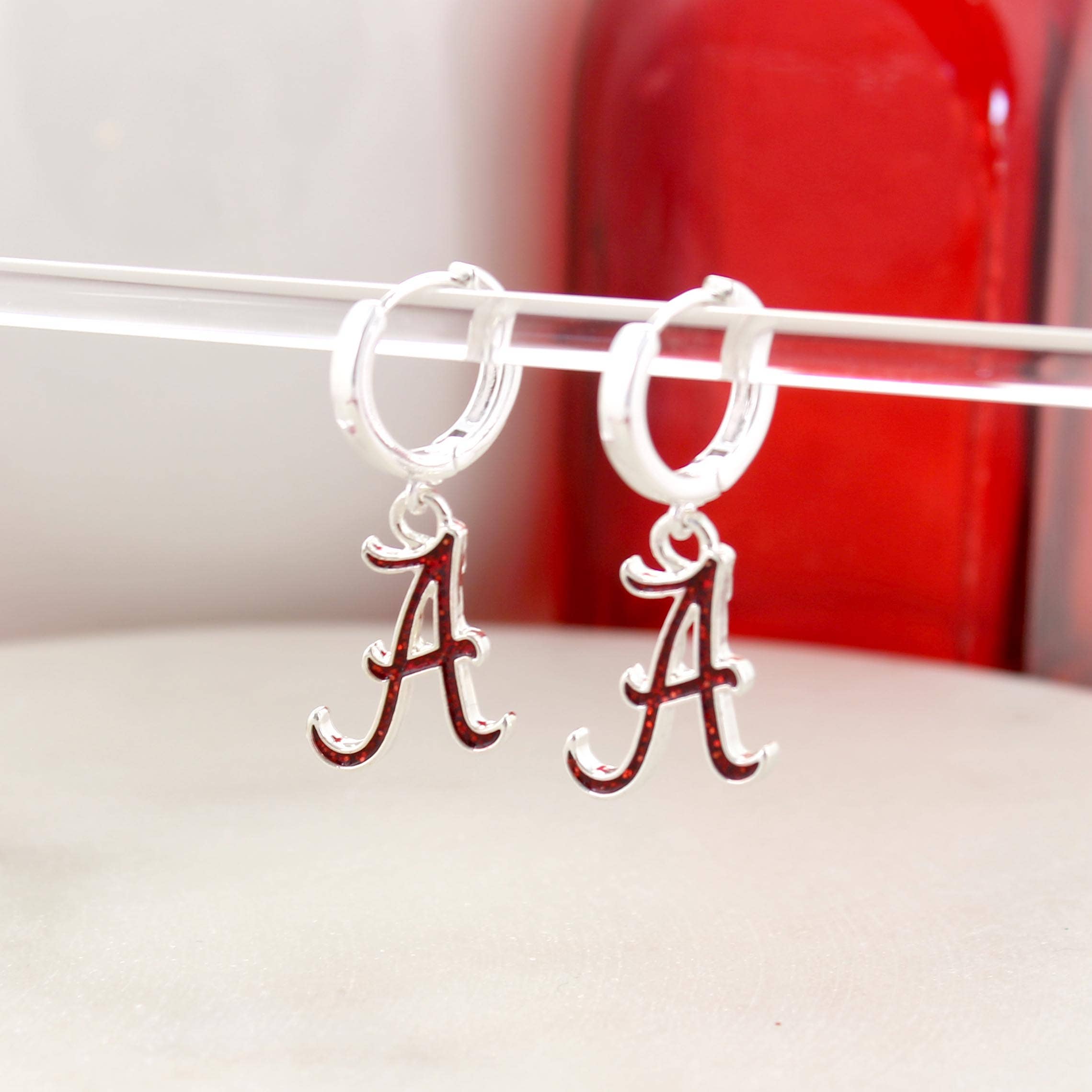 Seasons Jewelry Alabama Glitter Enamel Logo Huggie Hoop Earrings feature silver huggie hoops with dangling red and white Alabama “A” logo charms accented by crimson glitter enamel, shown on a clear rod with a red object in the background.