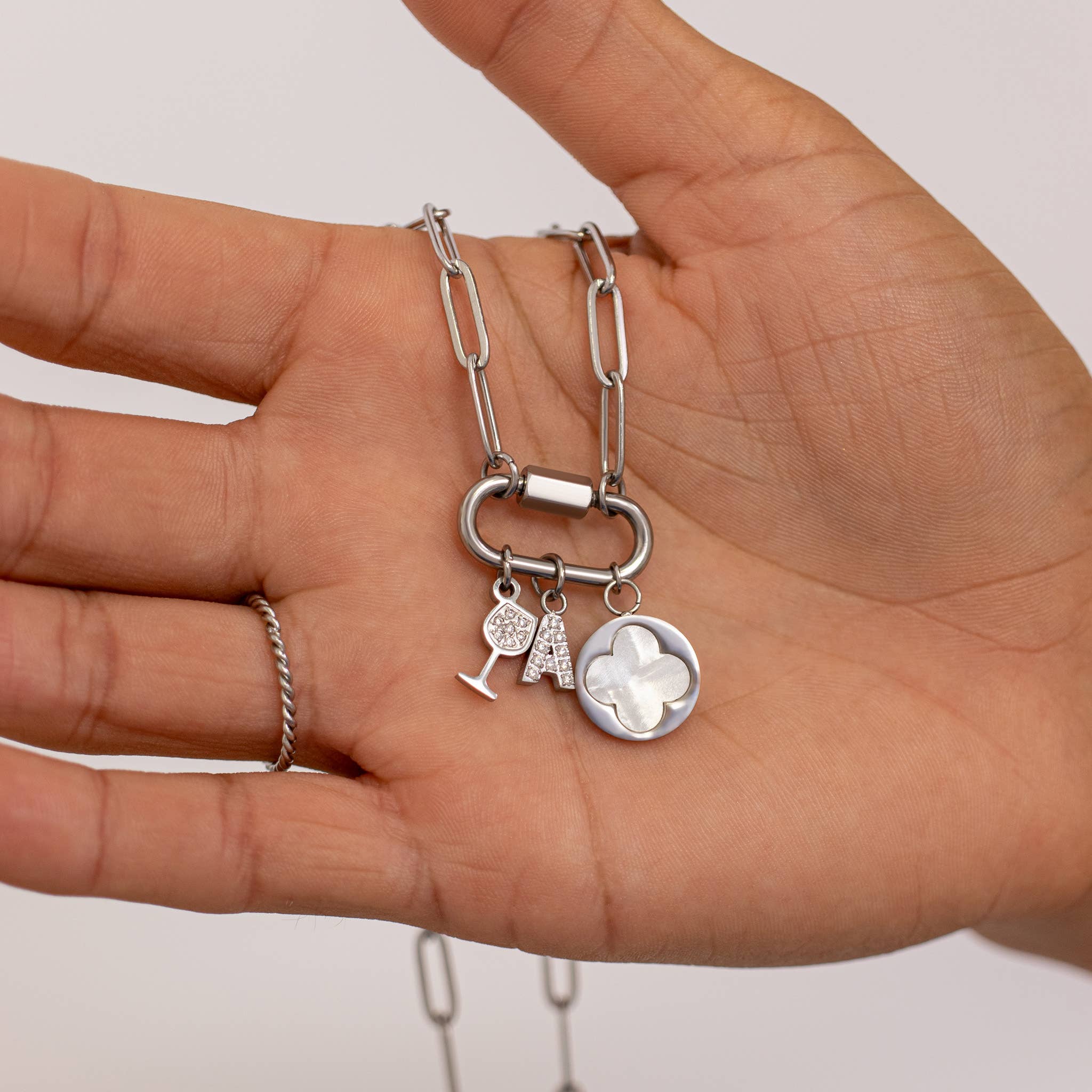 A hand holds the WJW 18K Gold PVD Paperclip Carabiner Necklace, which features three charms: a wine glass, a tennis racket with ball, and a round pendant adorned with a clover design.