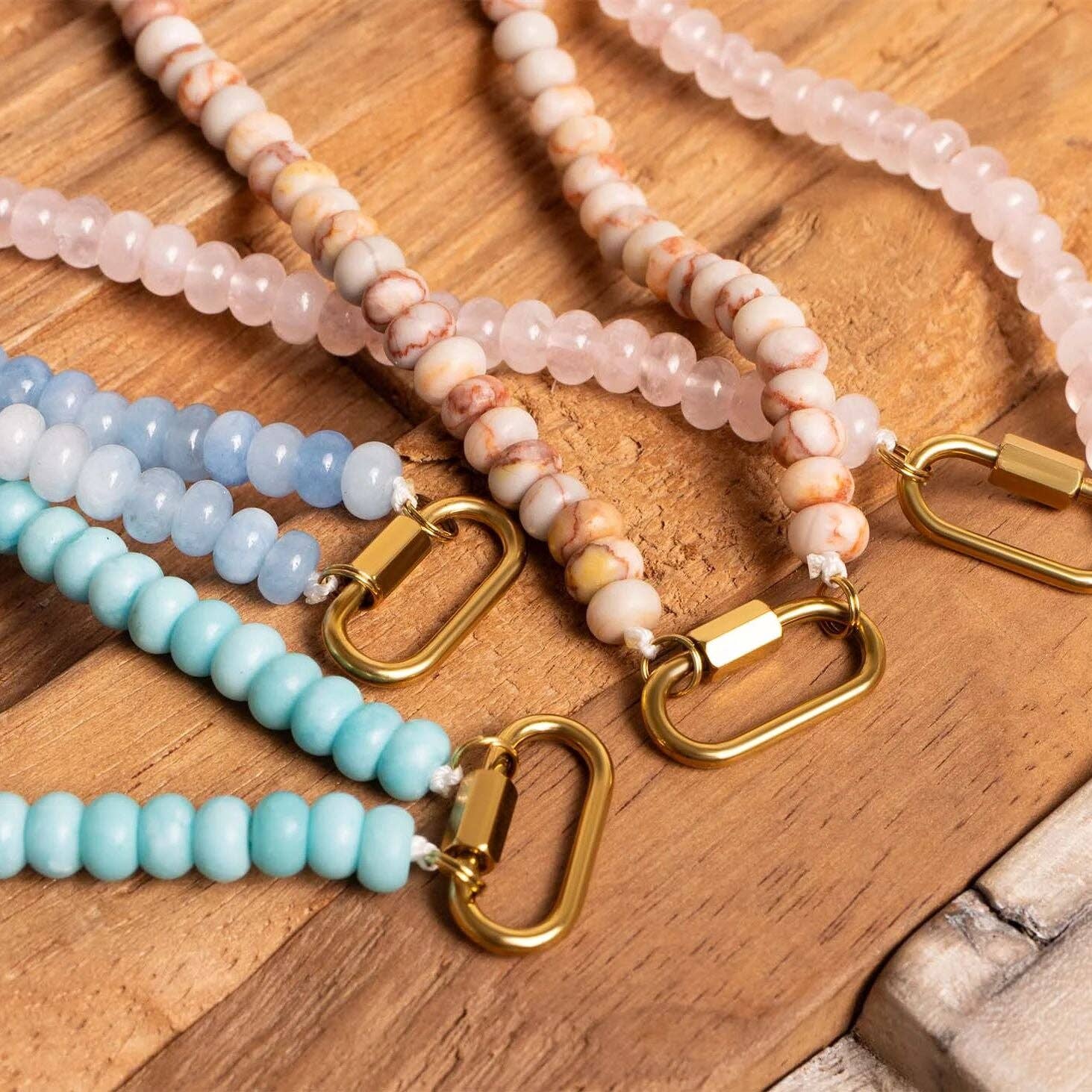 Four beaded keychains with gold clasps rest on a wooden surface, their beads in blue, pink, and cream shades reminiscent of the WJW Stainless Steel Turquoise Chunky Bead Carabiner Necklace, adding a vibrant pop of color.