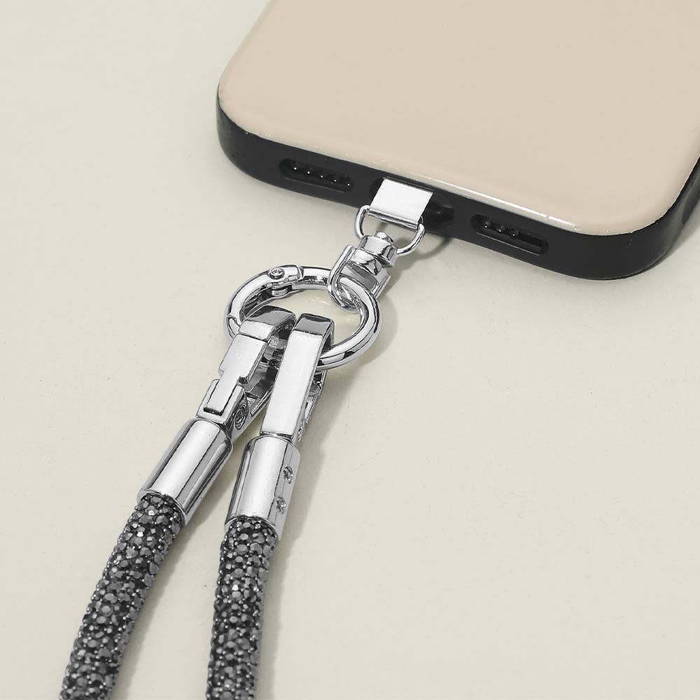 A close-up of a beige smartphone with a black edge, secured to the Sensibling Corp. Rhinestone Phone Lanyard Necklace—a gray and silver lanyard—via a metal clip and ring, resting on a light surface.