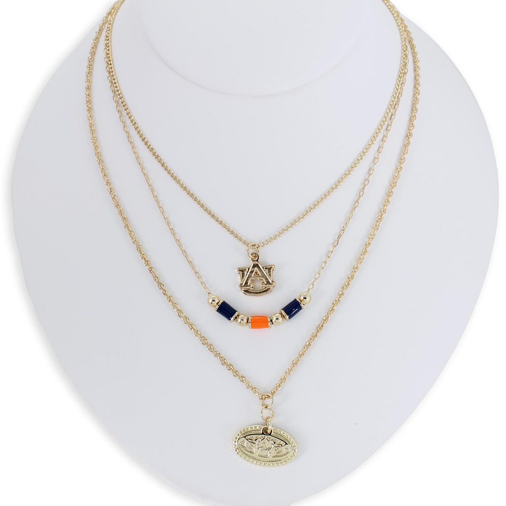 The Auburn Gold & Tube Bead Trio Necklace Set by Seasons Jewelry features three gold-tone layered necklaces displayed on a white bust, showcasing an AU logo, navy and orange tube bead accents, and an oval pendant with a raised design—an ideal War Eagle accessory for spirited Auburn University fans.