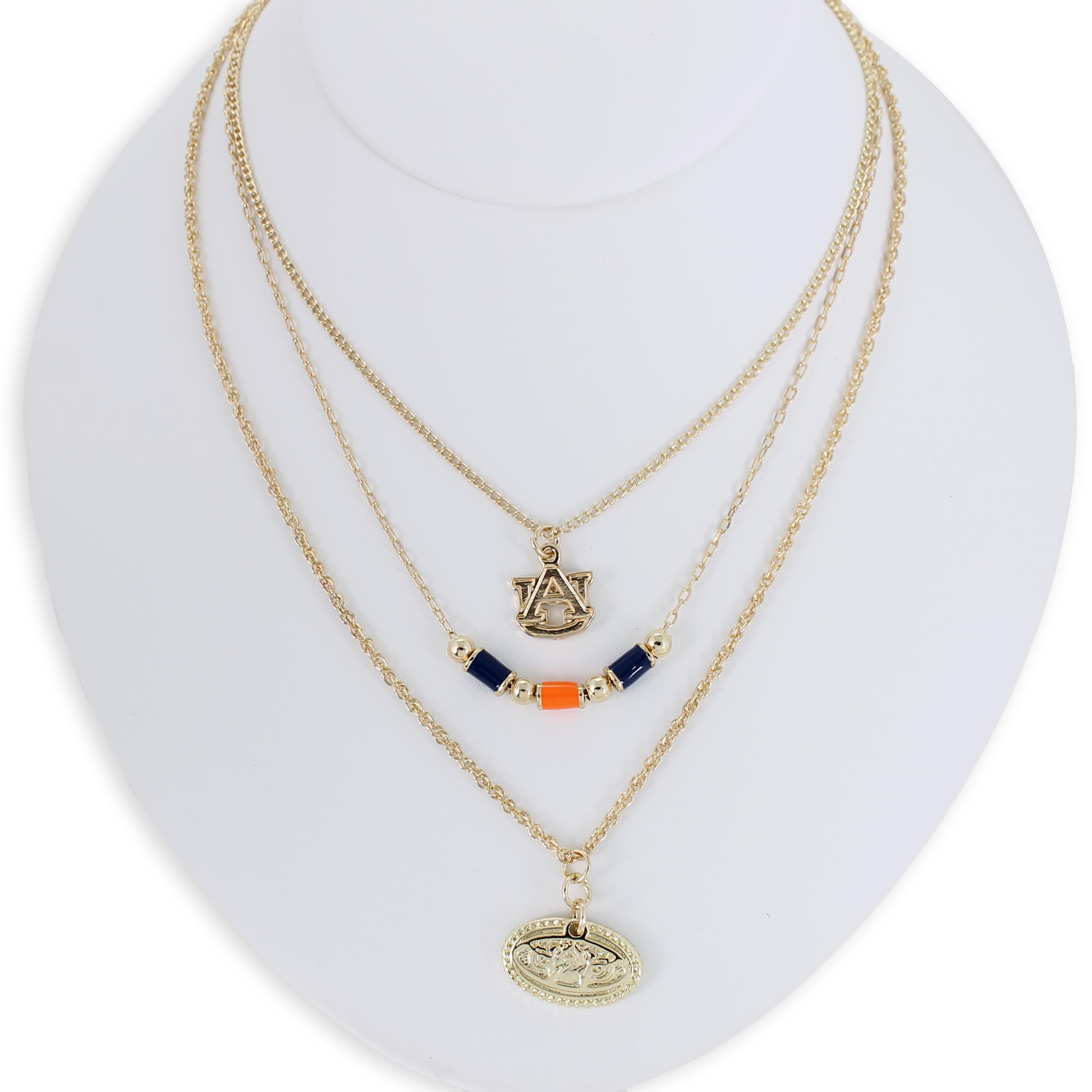 The Auburn Gold & Tube Bead Trio Necklace Set by Seasons Jewelry features three gold-tone layered necklaces displayed on a white bust, showcasing an AU logo, navy and orange tube bead accents, and an oval pendant with a raised design—an ideal War Eagle accessory for spirited Auburn University fans.