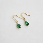 A pair of Ellemadera Chic Gemstone Dangle Earrings, featuring teardrop-shaped green gemstones and hypoallergenic gold settings, displayed on a white fabric background.