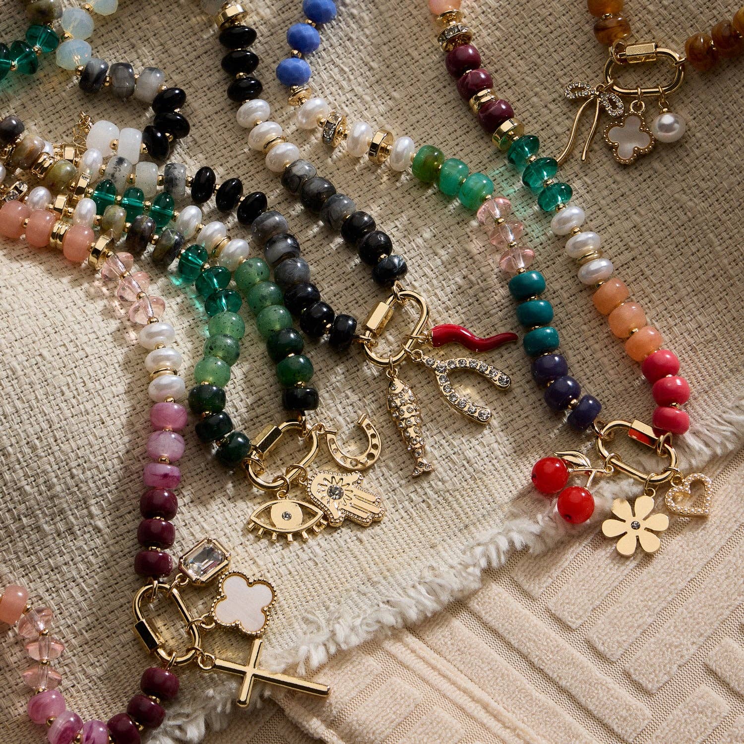 Lilla Haven’s Textured Glass Bead Necklace with Charm Trio is beautifully displayed on textured beige fabric. Each necklace features colorful glass beads and a trio of gold and enamel charms—such as flowers, hearts, eyes, cherries, or crosses—accented by gold-toned spacers that lend an elegant touch to each playful design. Main image