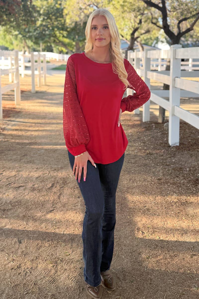 A person with long blonde hair stands on a dirt path by a white fence, wearing the Sequin Sleeve Blouse from 24-Hrs Fashion and dark jeans, with trees visible in the background.
