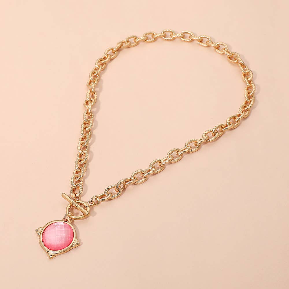 The Sensibling Corp. Faceted Round Stone Charm Pendant Toggle Necklace, featuring a gold chain with a pink round charm and toggle-style closure, is displayed on a light beige background.