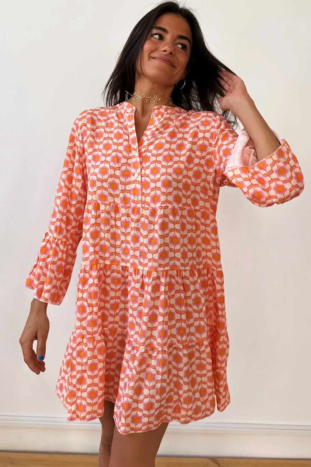 A woman in a Shewin Orange Floral Button V Neck Bell Sleeve Mini Dress stands against a plain background, smiling and gently touching her hair.