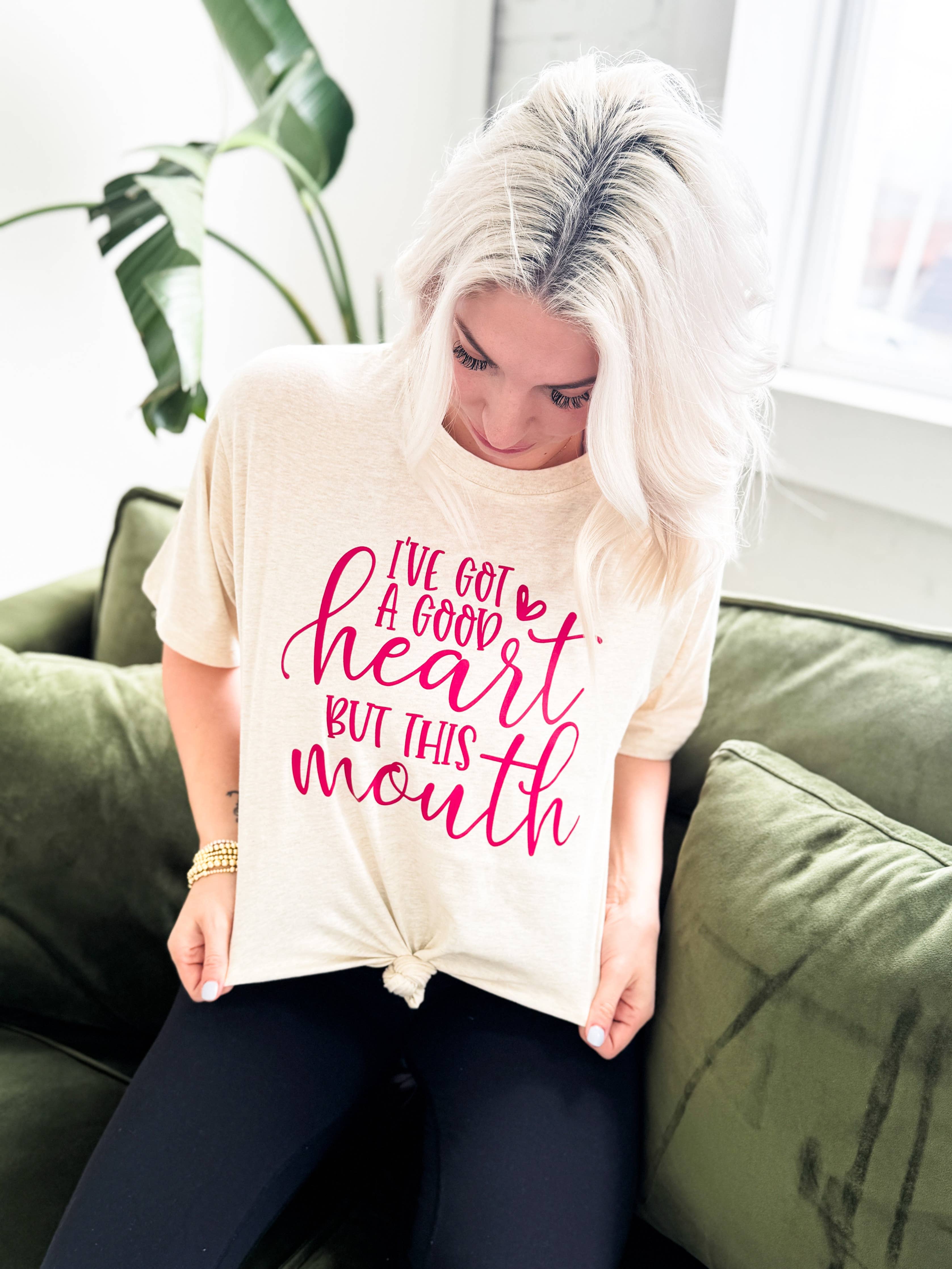 A woman with platinum blonde hair sits on a green couch, looking down at her Festivitees "I've Got a Good Heart But This Mouth" Graphic Tee, a beige unisex shirt with the phrase printed in pink lettering.