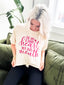 A woman with platinum blonde hair sits on a green couch, looking down at her Festivitees "I've Got a Good Heart But This Mouth" Graphic Tee, a beige unisex shirt with the phrase printed in pink lettering.