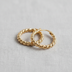 A pair of Ellemadera Sculptural Twisted Hoop Earrings, featuring small gold hoops with silver posts, are displayed on a light textured fabric background.