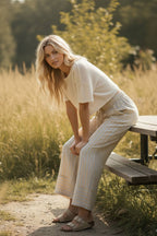 A woman with long blond hair sits on a wooden bench in a grassy field, wearing Sewn and Seen’s Woven Striped Wide Leg Pants, a white top, and sandals.