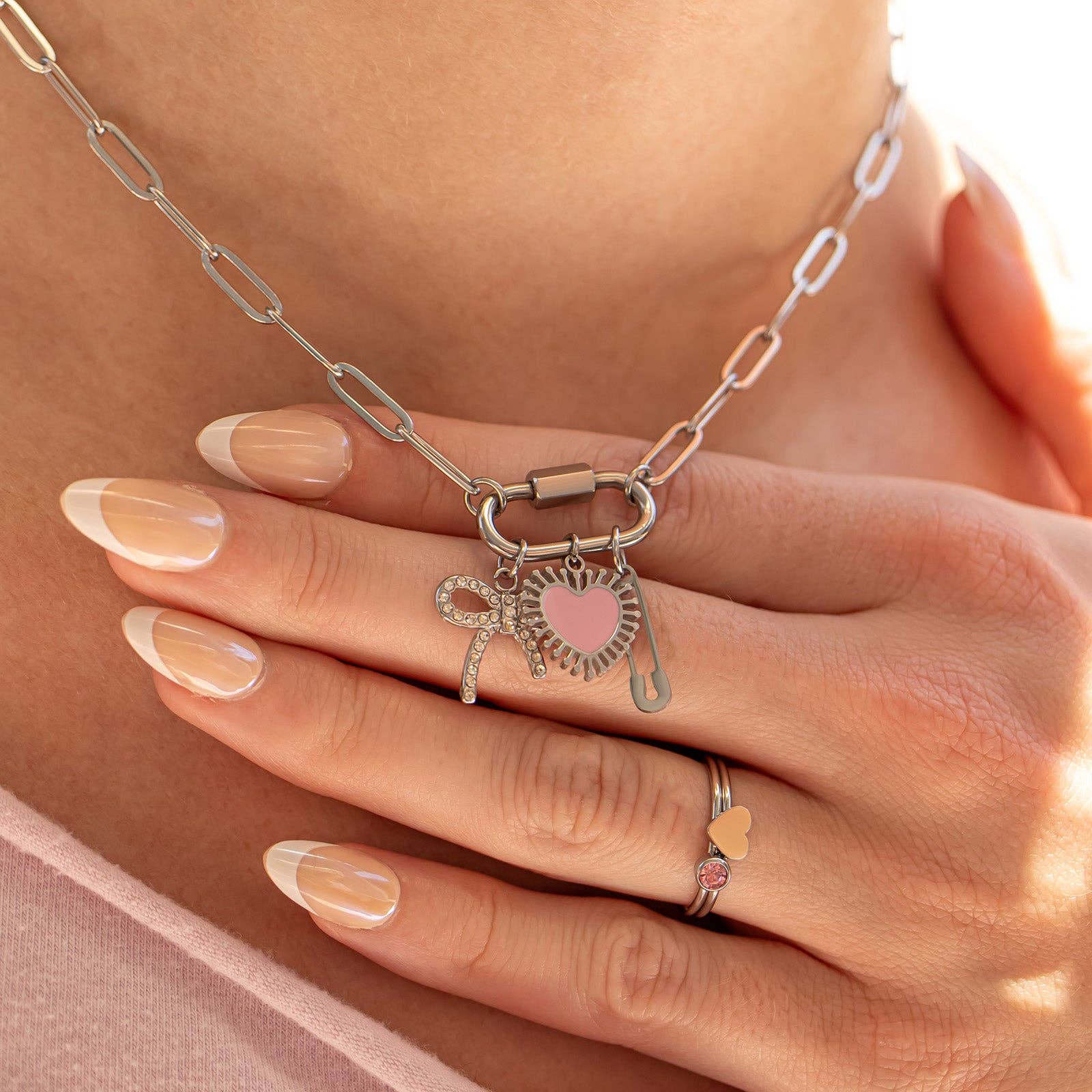 A hand with manicured nails rests on a chest, showcasing the WJW 18K Gold PVD Paperclip Carabiner Necklace—a customizable piece featuring a paperclip chain adorned with heart, lock, and key charms—paired with a matching ring accented by a pink heart detail.