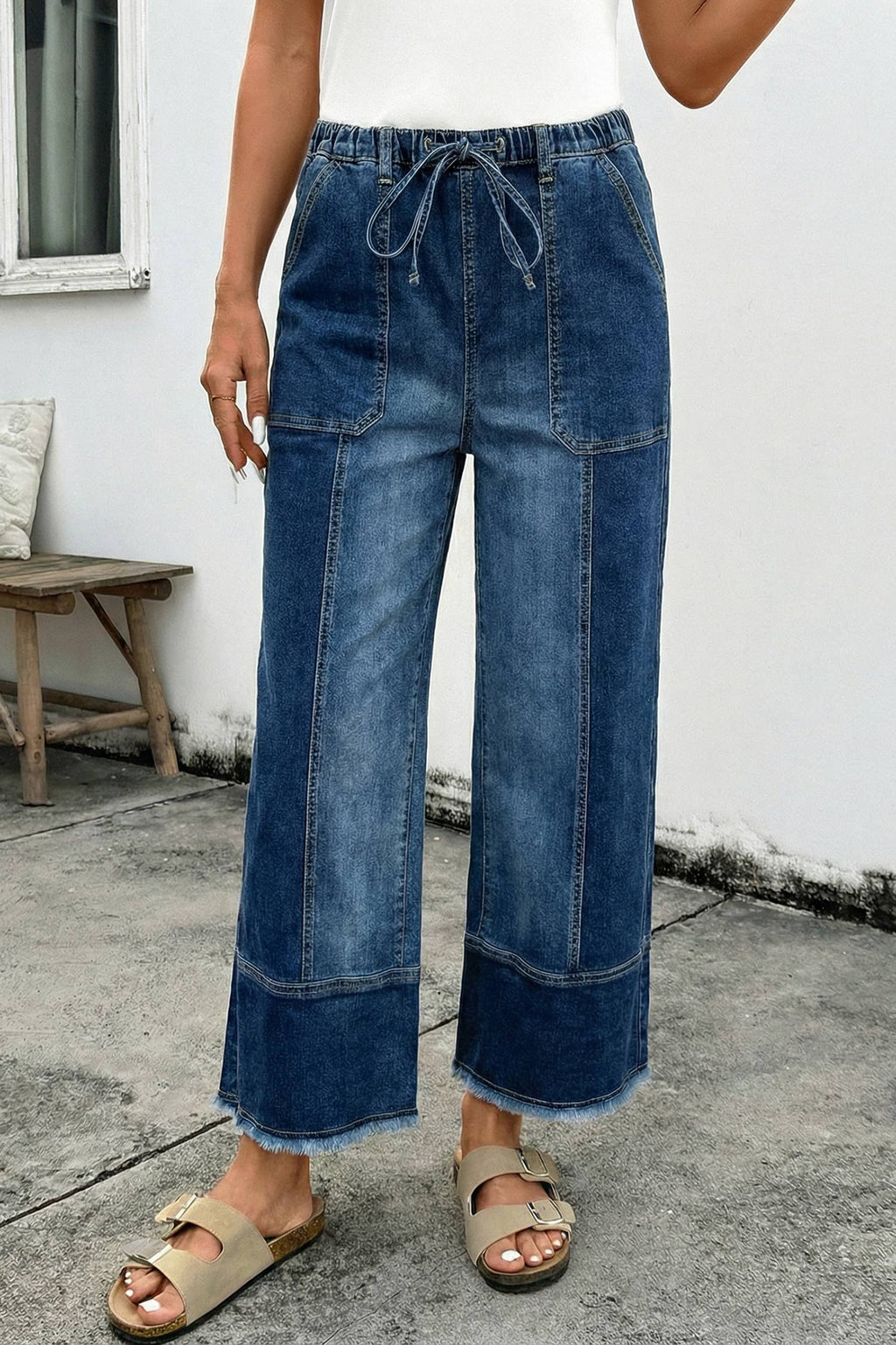 A person in 1 the Woman Drawstring Seam Detail Raw Hem Wide Leg Denim Pants, beige sandals, and a white top stands on concrete by a bench against a white wall.