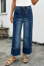 A person in 1 the Woman Drawstring Seam Detail Raw Hem Wide Leg Denim Pants, beige sandals, and a white top stands on concrete by a bench against a white wall.