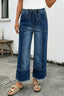 A person in 1 the Woman Drawstring Seam Detail Raw Hem Wide Leg Denim Pants, beige sandals, and a white top stands on concrete by a bench against a white wall.