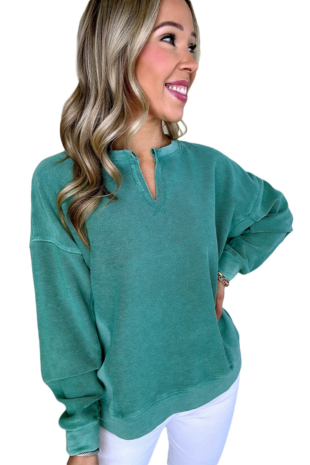 A smiling person poses with one hand on their hip, wearing a Notched Neck Exposed Seam Drop Shoulder Sweatshirt in green by Shewin and white pants.