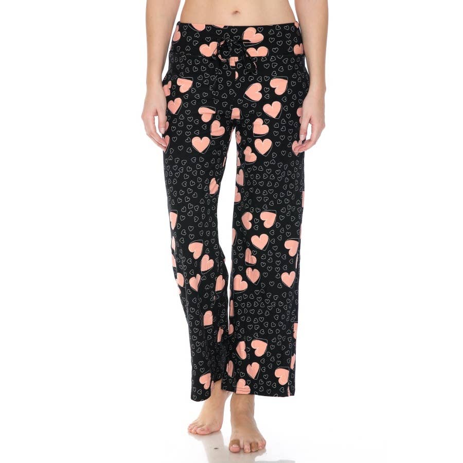 A person stands barefoot against a white background, wearing Leggings Depot Buttery Soft Pajama Pants in black with pink hearts and white swirl patterns, featuring an elastic waistband.
