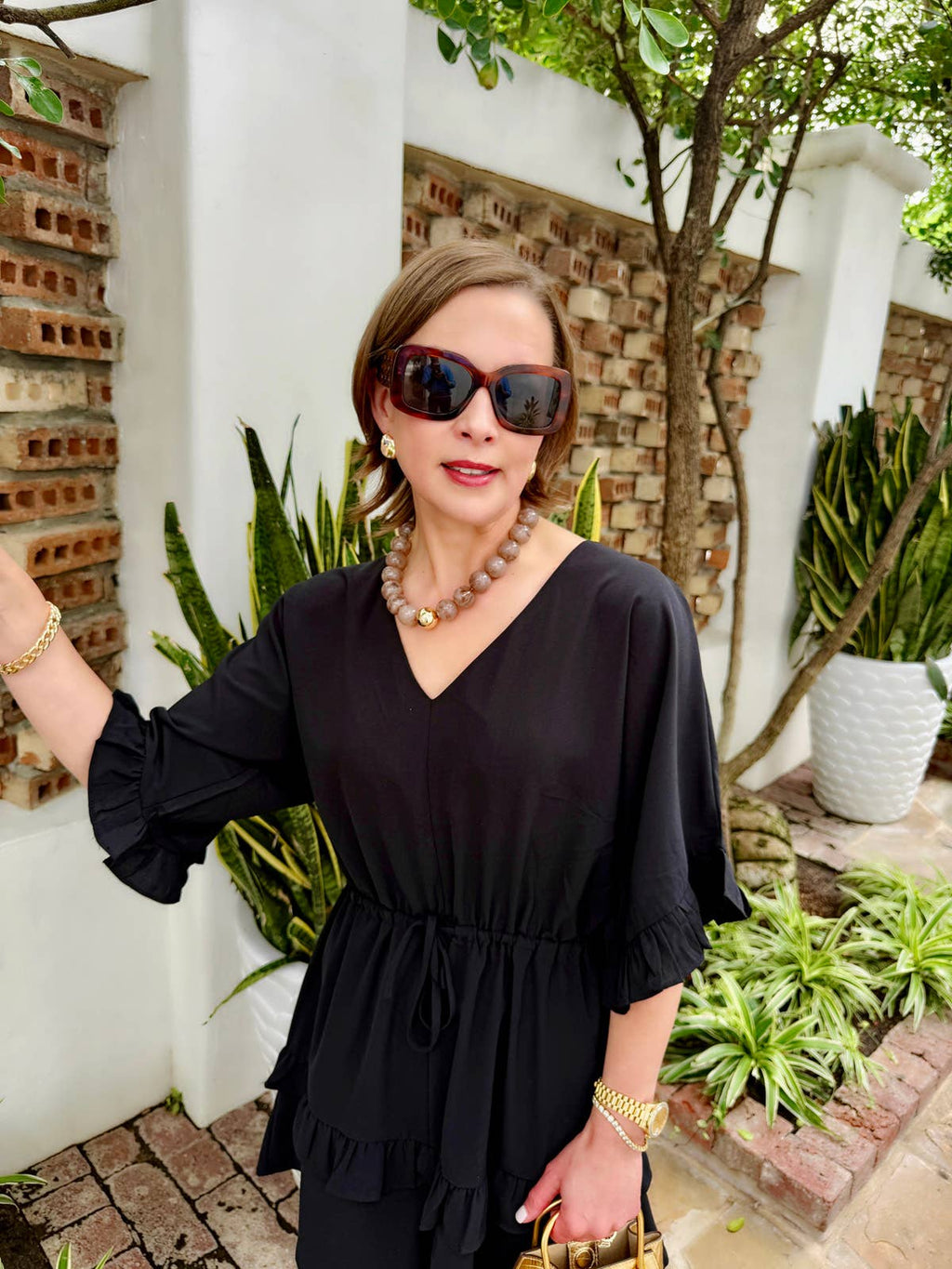 A woman wearing a black blouse, sunglasses, and jewelry—including the Marjorie Fall Beaded Necklace by Canvas Style, featuring oversized resin beads—stands outdoors near plants and a white wall with brick accents.