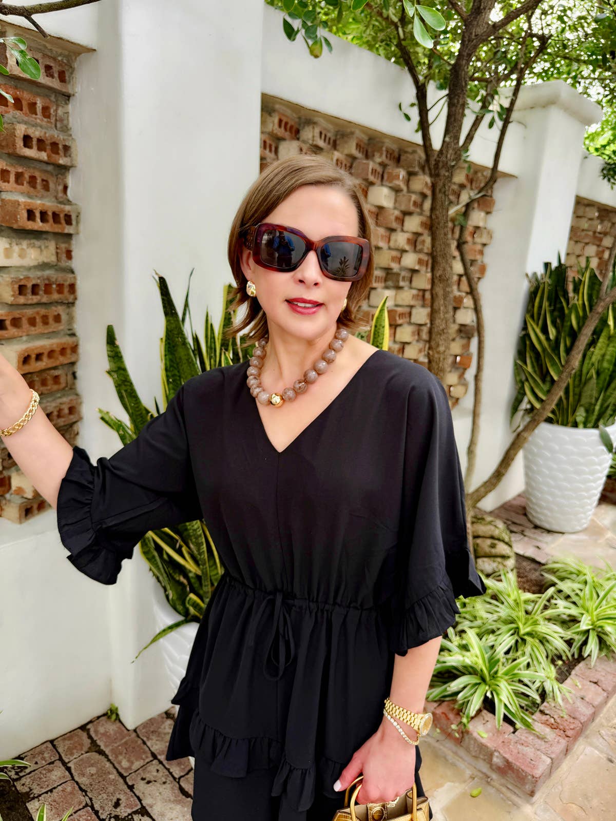 A woman wearing a black blouse, sunglasses, and jewelry—including the Marjorie Fall Beaded Necklace by Canvas Style, featuring oversized resin beads—stands outdoors near plants and a white wall with brick accents.