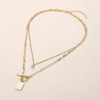 The Sensibling Corp. Stainless Steel Mother of Pearl Rectangular Frame Toggle Pendant Round Stone Accent Layered Necklace features two elegant chains: one with a rectangular mother of pearl pendant framed in stainless steel and secured with a chic toggle clasp, the other adorned with a single small round stone accent. Displayed on a light beige background, this modern necklace beautifully combines style and durability.