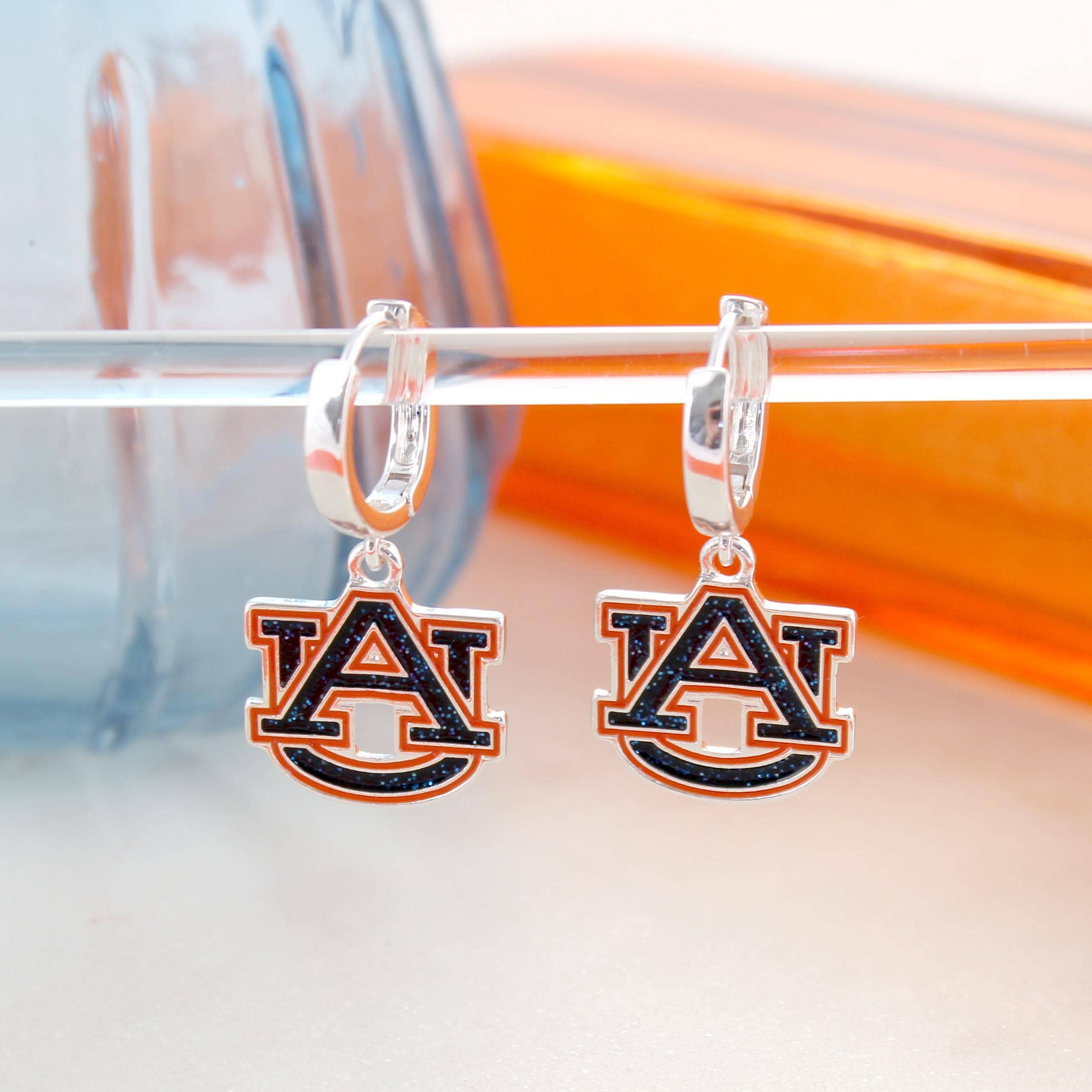 A pair of Auburn Glitter Enamel Logo Huggie Hoop Earrings by Seasons Jewelry, featuring sparkling "AU" logo charms, perfect for game day, displayed on a clear rod with glass objects in the background.