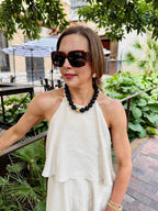 A woman stands outdoors near greenery and a patio area wearing a sleeveless white dress, large sunglasses, and the Canvas Style Marjorie Game Day Beaded Necklace.
