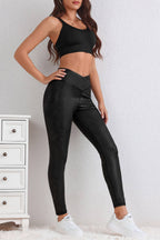 A woman stands indoors wearing LA Society's Black Crossed Dip Waist Sleek Leather Leggings with a foil-embossed finish, a black sports bra, and white sneakers beside a small white dresser.