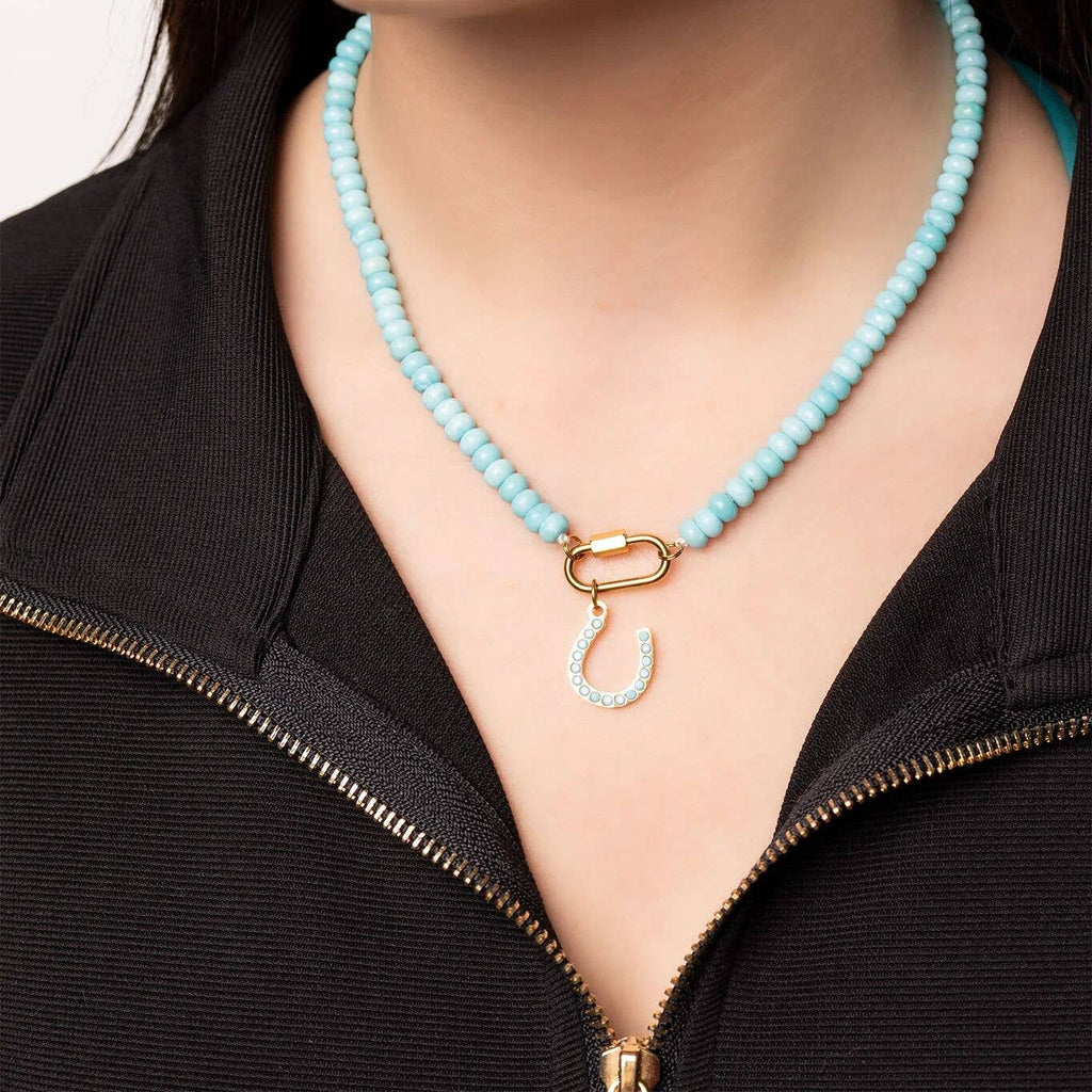 A person wearing the WJW Stainless Steel Turquoise Chunky Bead Carabiner Necklace, featuring vibrant turquoise beads, a secure gold-tone carabiner clasp, and a distinctive horseshoe-shaped pendant, styled over a partially unzipped black top. This necklace artfully combines bold style with the lasting durability of stainless steel.