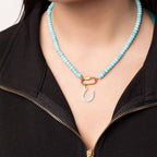 A person wearing the WJW Stainless Steel Turquoise Chunky Bead Carabiner Necklace, featuring vibrant turquoise beads, a secure gold-tone carabiner clasp, and a distinctive horseshoe-shaped pendant, styled over a partially unzipped black top. This necklace artfully combines bold style with the lasting durability of stainless steel.
