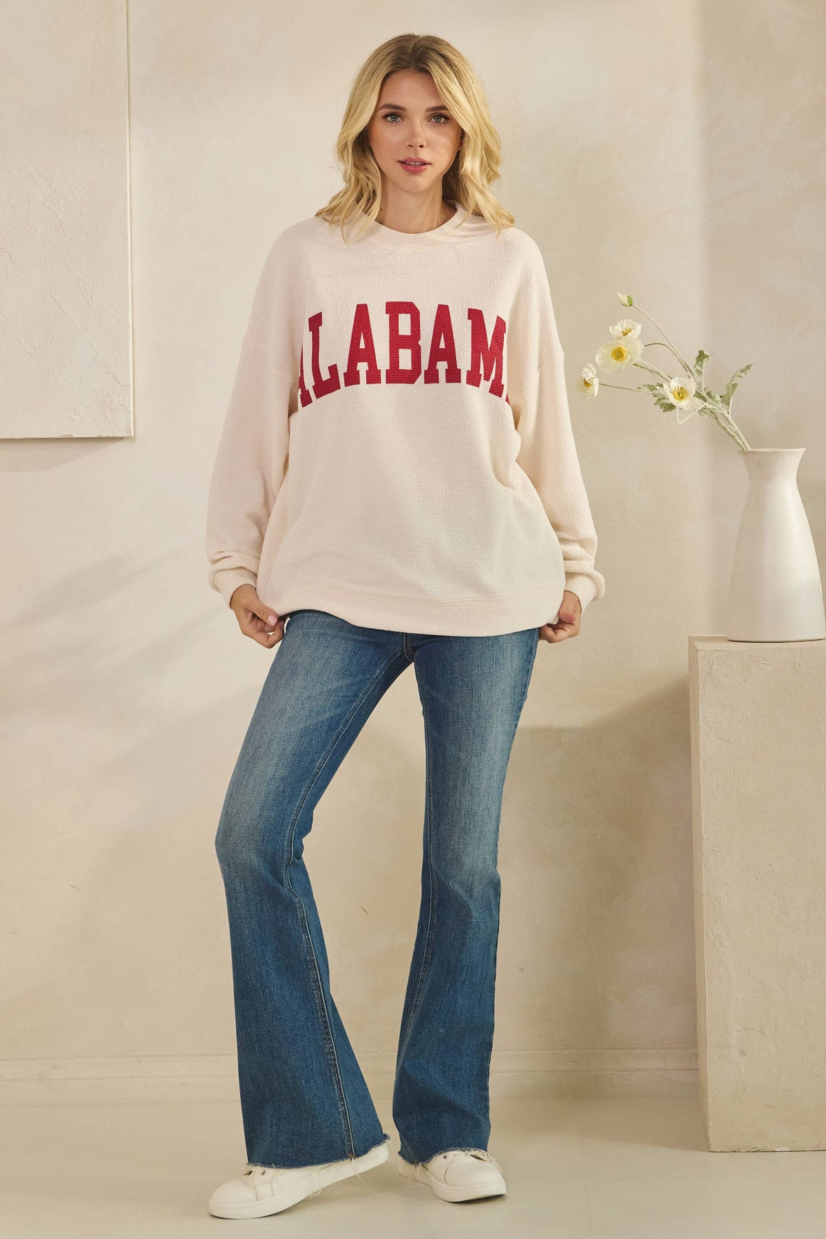 A woman wears a cream-colored ALABAMA Cloudy Soft Knit Long Sleeve Top with red lettering, paired with blue flared jeans and white sneakers.