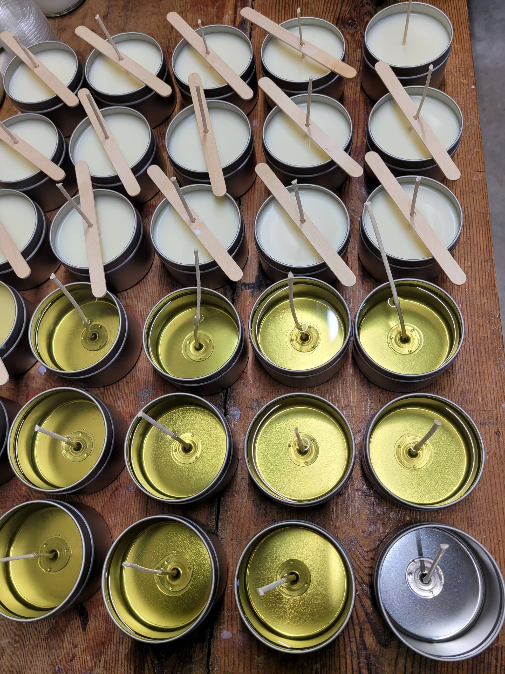 A wooden table displays a collection of 4oz Mahogany Teakwood soy wax candles in tins by Stephanie Henry Candle Company. Some are white and unset, others yellow and set, with popsicle sticks across them, ready to support wood wicks for an inviting fragrance when lit.