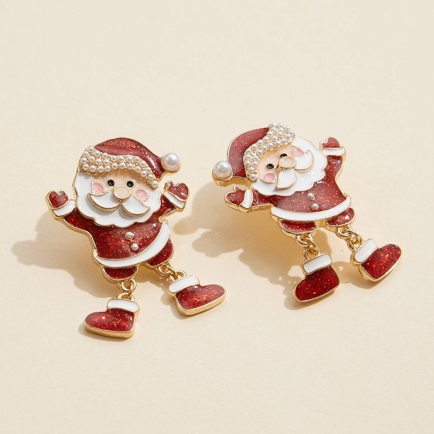 The Enamel Glitter Santa Dangle Earrings from Lilla Haven feature a festive Santa Claus design with red and white enamel, gold outlining, and pearl accents. Displayed on a beige background, these Pearl Holiday Statement Earrings are the perfect accessory to brighten up your seasonal look.