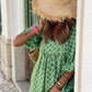 A person in a Rosa Clothing Bohemian Floral Balloon Sleeves loose flare swing dress and a large straw hat leans against a white wall, partially covering their face with the hat.