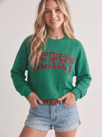 Woman wearing a Faire Merry Plaid Cloudy Knit Top with “MERRY” in red and black plaid lettering across the chest and blue denim shorts.