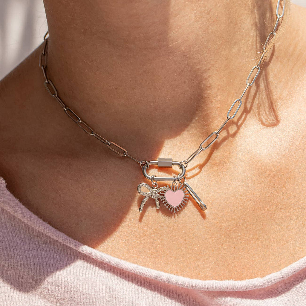 A person wearing the WJW 18K Gold PVD Paperclip Carabiner Necklace, adorned with charms such as a pink heart, a rhinestone bow, a wing, and a wand.